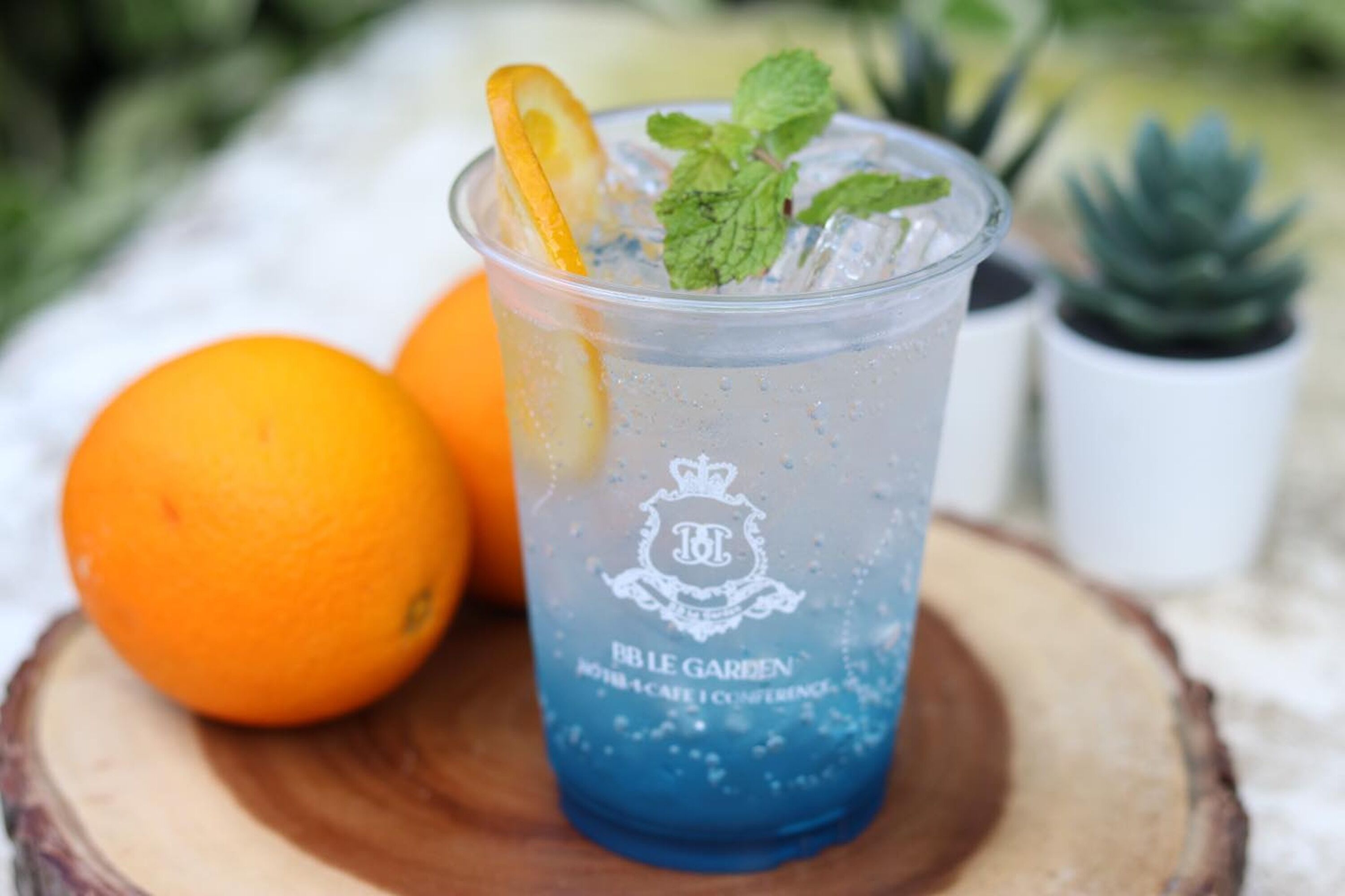 A clear glass with a blue liquid, ice, a slice of orange, and mint leaves, labeled "BB Le Jardin."