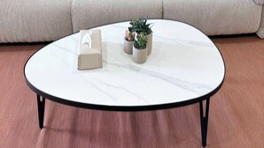 Uma mesa redonda branca com pernas pretas, um sofá bege com almofadas e uma pequena planta em vaso.