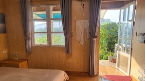 A wooden room with a bed, a small table with bottles, and a view through a window to greenery outside. - Books Home Dalat (Xuan Huong - Da Lat)
