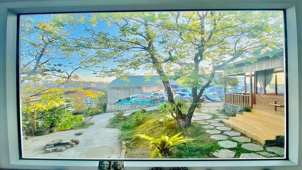 A view through a window of a garden with a stone pathway, a wooden deck, and a building with a green roof in the background. - Books Home Dalat (Xuan Huong - Da Lat)