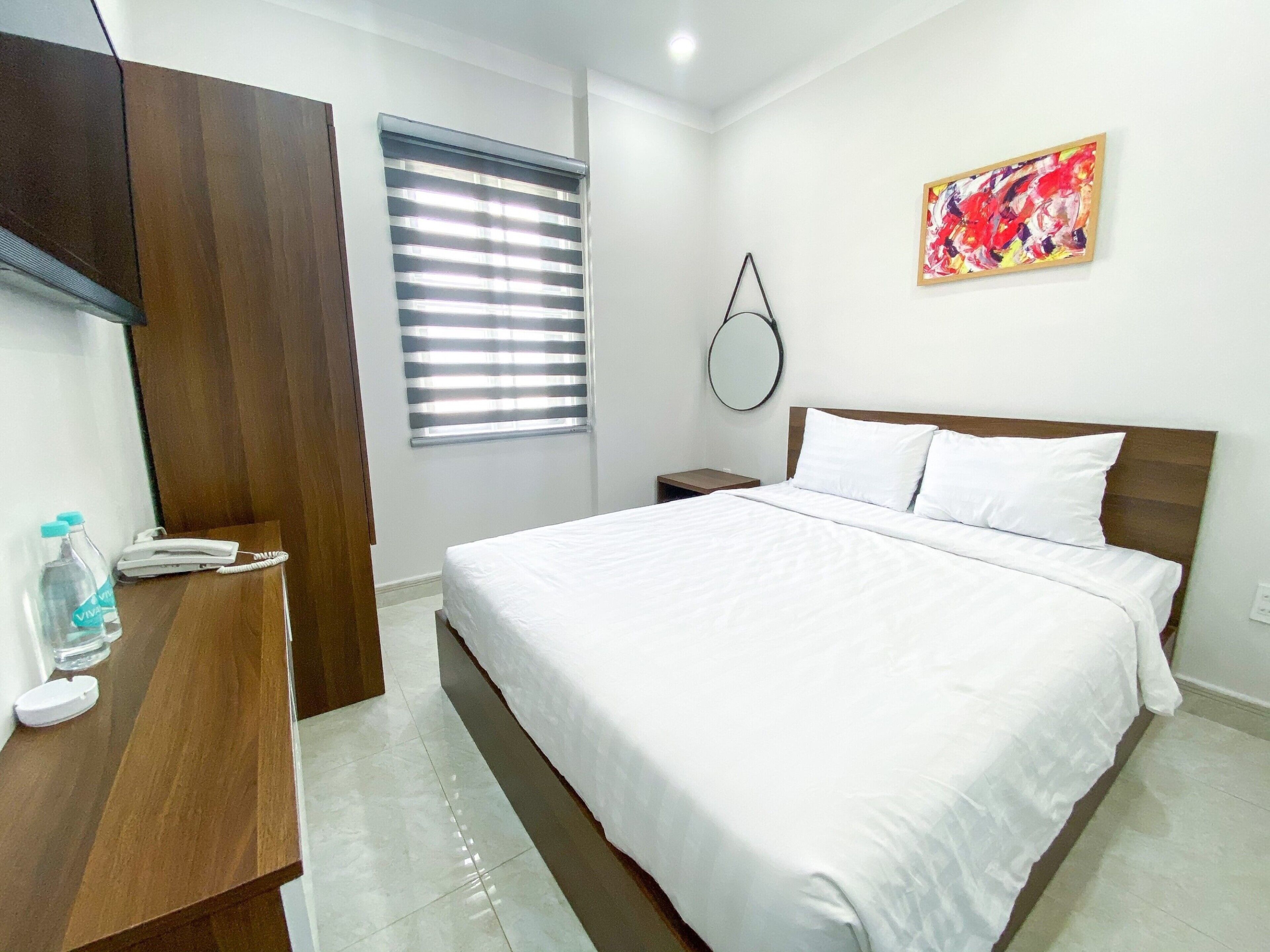 A hotel room with a bed, a wooden desk, a telephone, a water bottle, a mirror, and a painting on the wall.