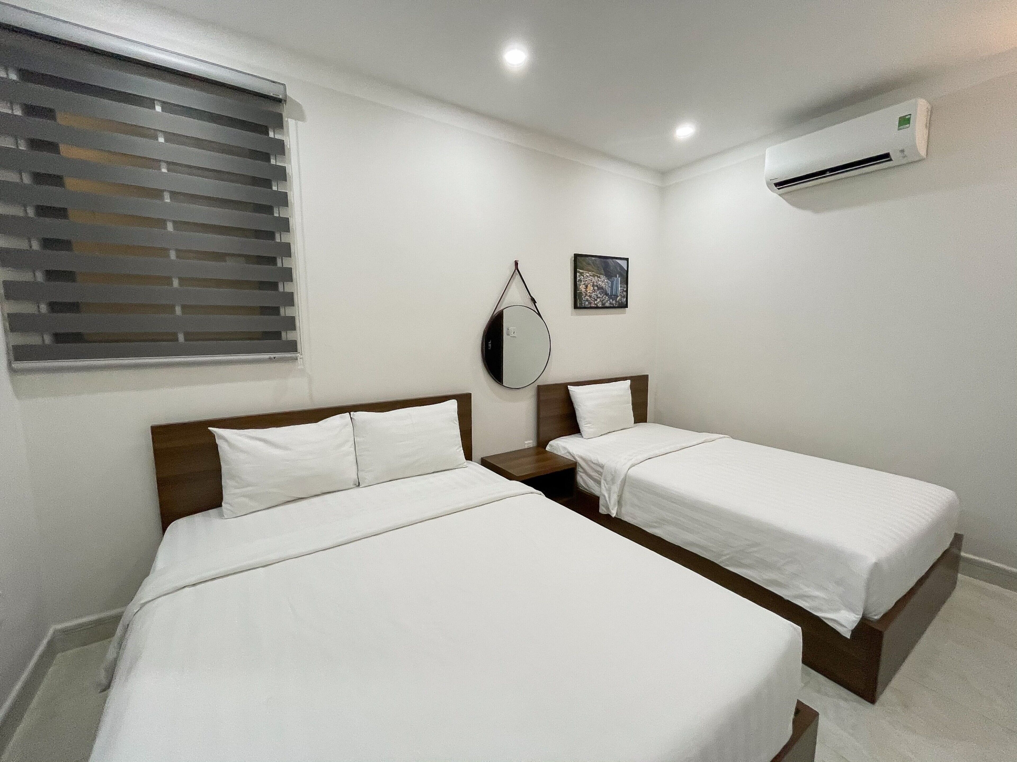 A hotel room with two single beds, a bedside table, a mirror, a window with blinds, and an air conditioning unit.