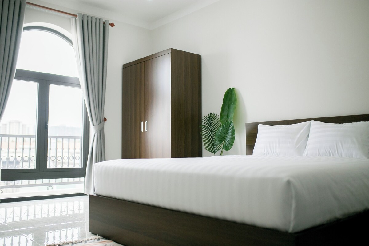 A hotel room with a large bed, a wardrobe, a window with curtains, and a plant.