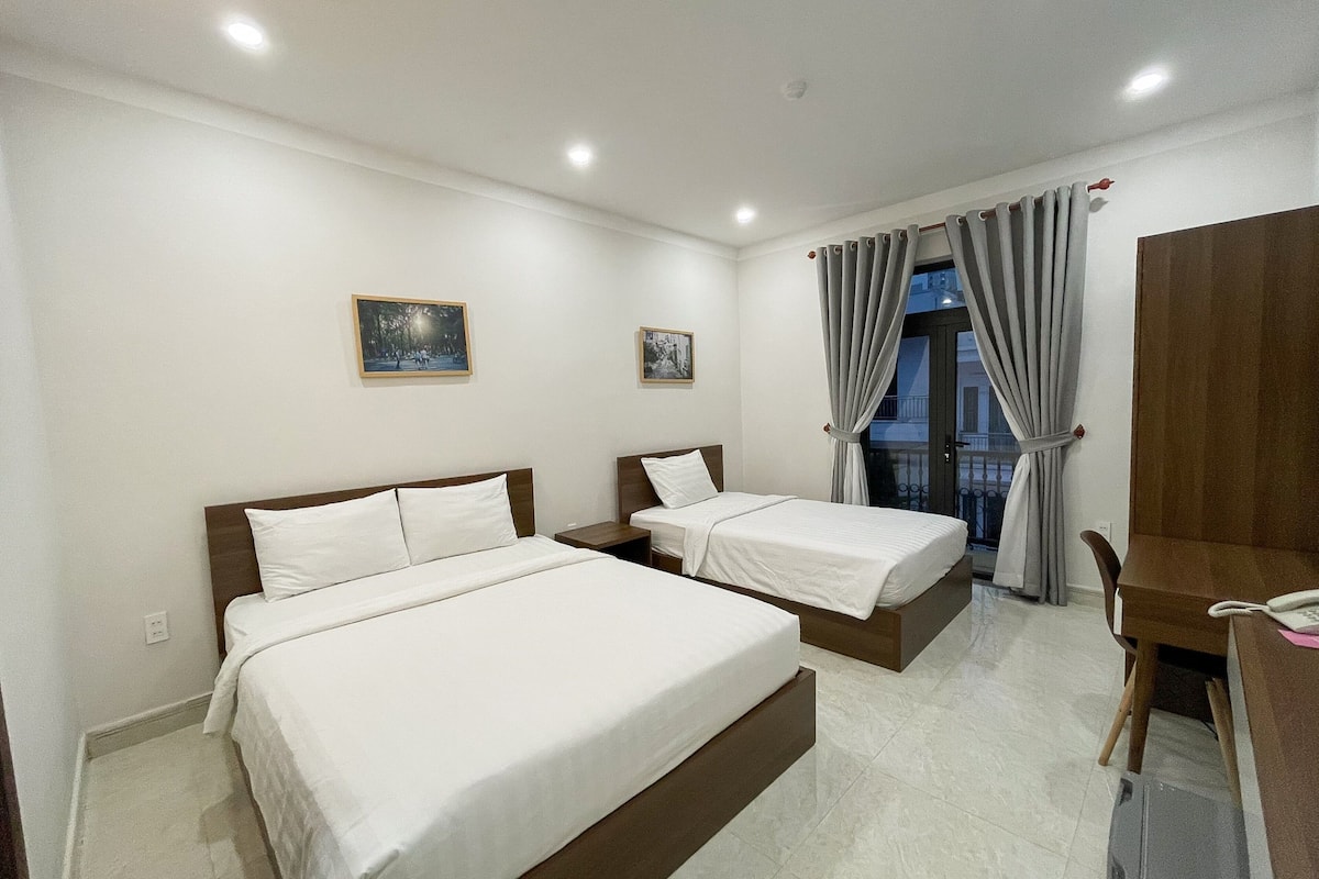 A hotel room with two beds, a desk, and a chair. There are two paintings on the wall and a window with curtains.