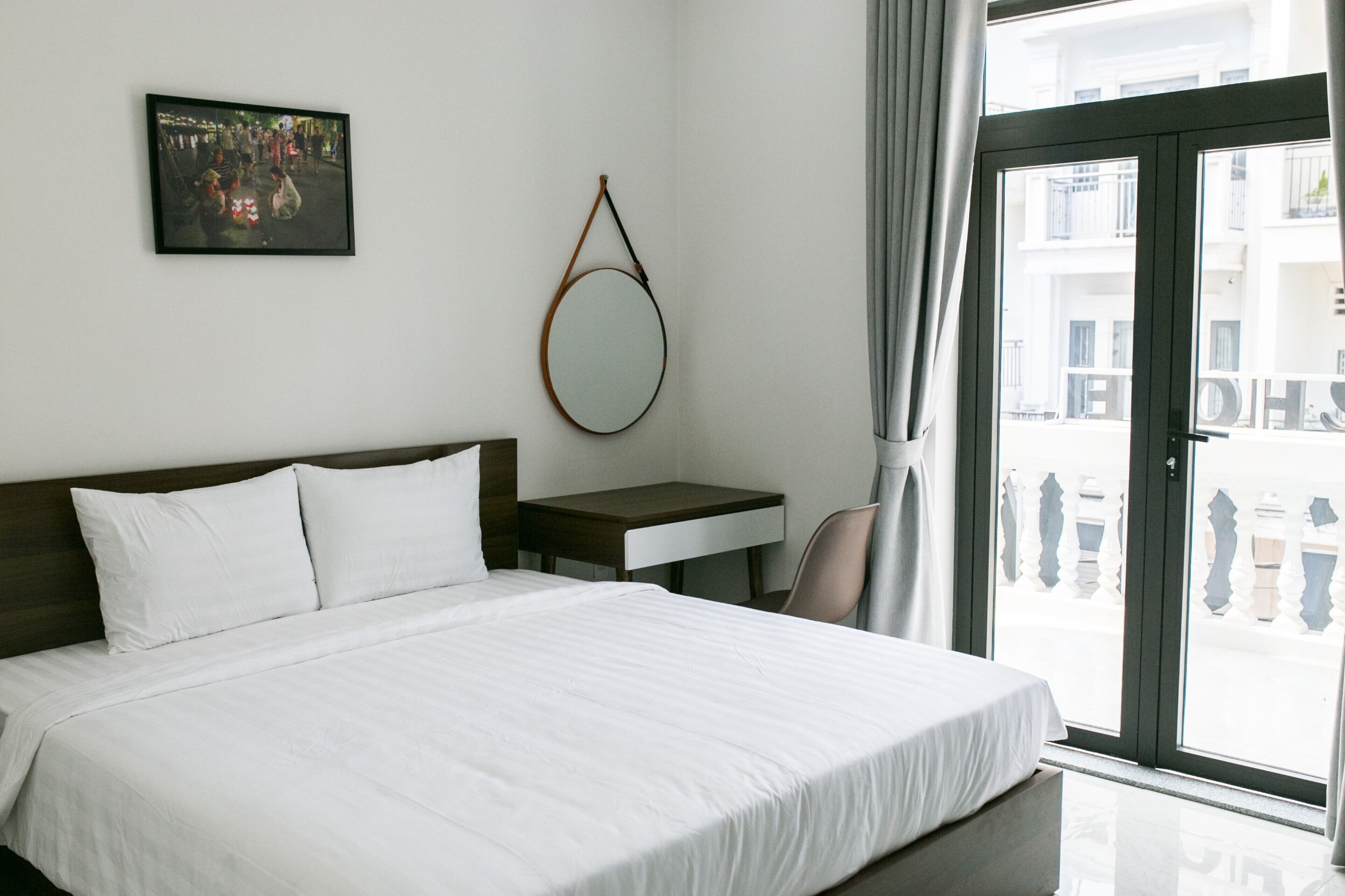 A hotel room with a bed, a desk, a chair, a round mirror, and a balcony with a railing.