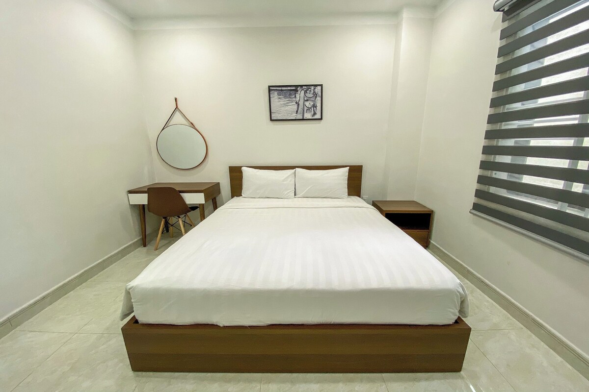 A bedroom with a bed, a desk, a chair, a nightstand, a mirror, and a window with blinds.