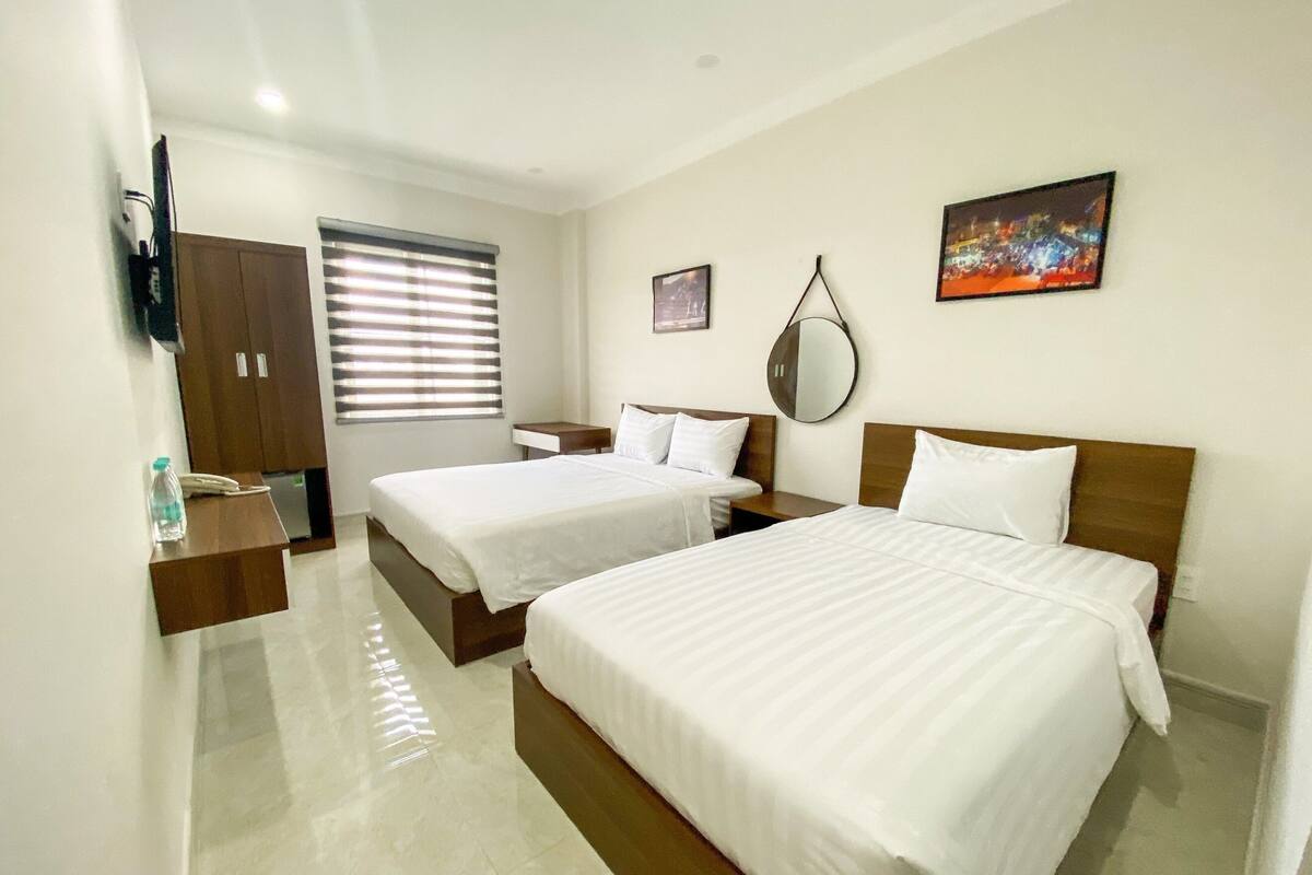 A hotel room with two beds, a wooden wardrobe, a bedside table with a lamp, a large mirror, and a painting on the wall.