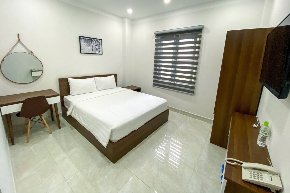 A hotel room with a bed, a desk, a chair, a mirror, a window with blinds, a telephone, and a water bottle.