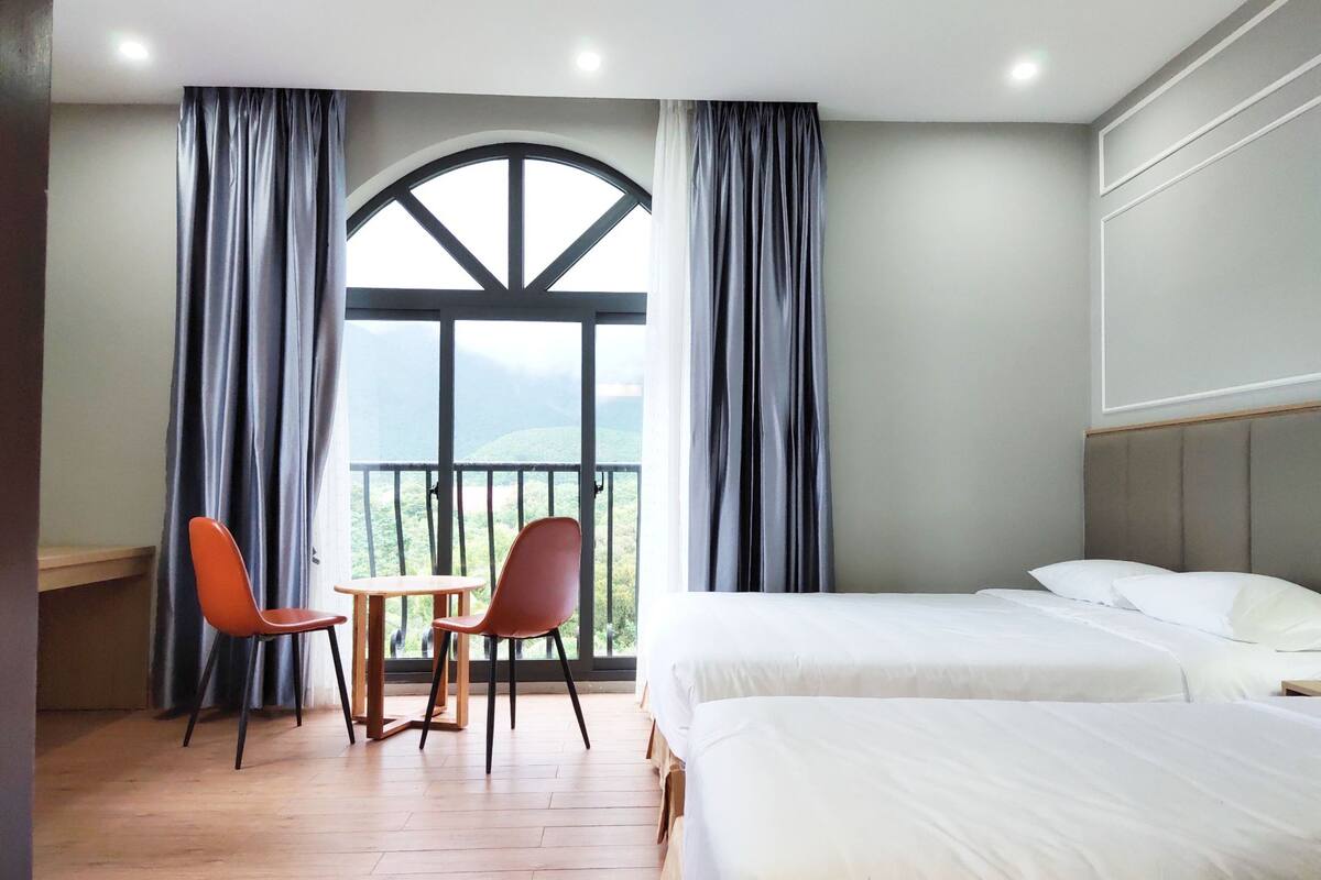 A hotel room with two beds, a balcony with curtains, and a table with chairs.