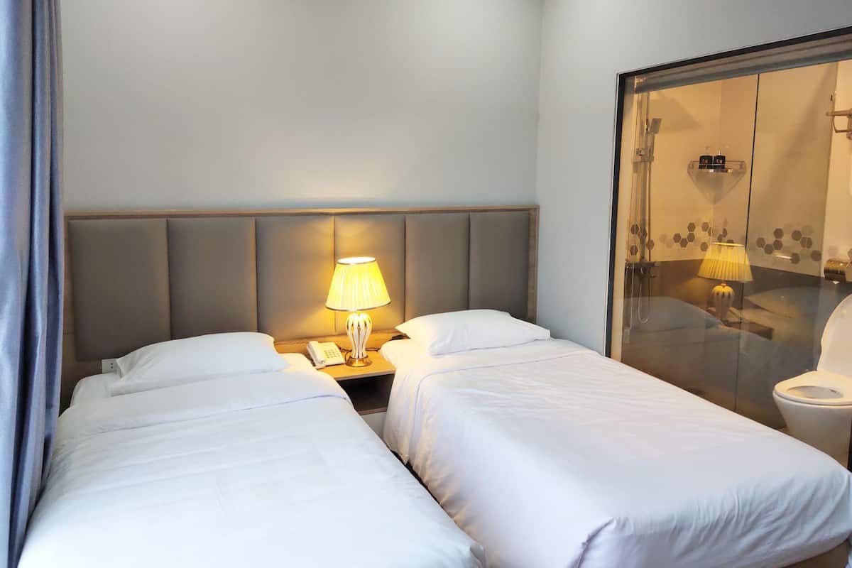 A hotel room with two beds, a nightstand with a lamp, a glass shower enclosure, and a toilet.