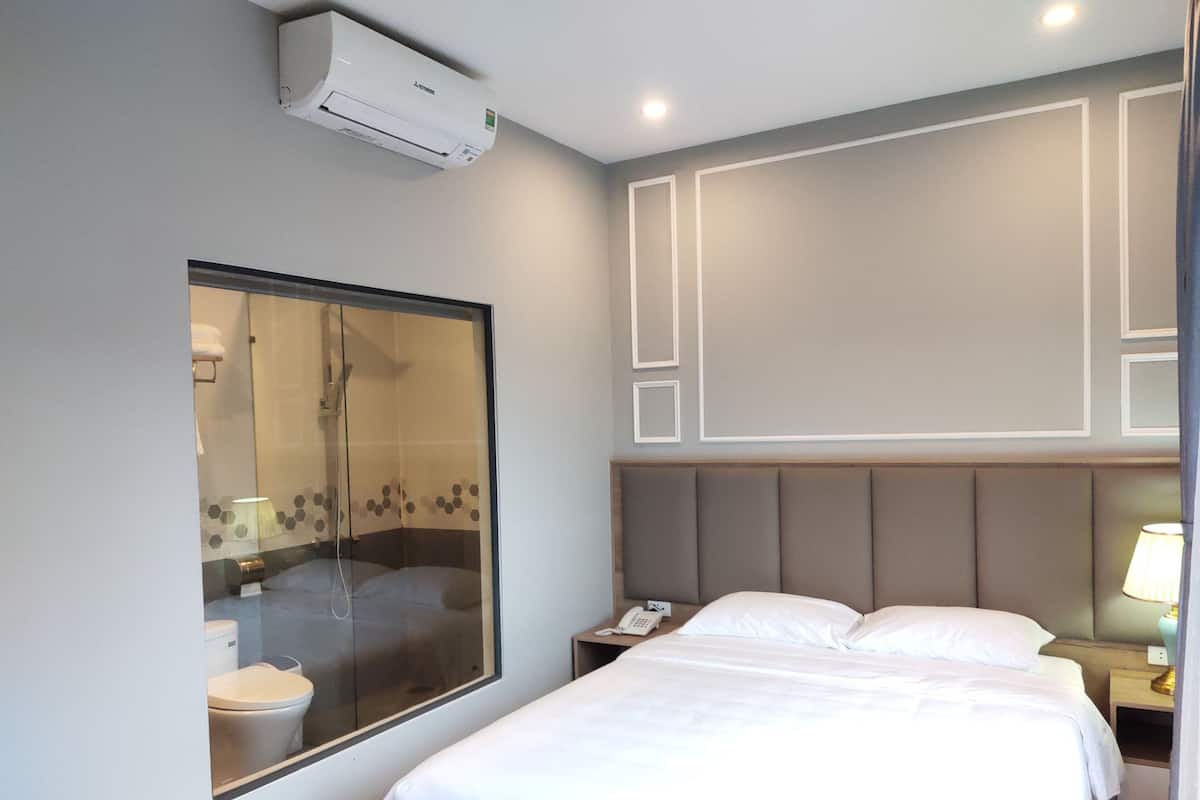 A hotel room with a large window, a bed, a bedside table with a lamp, and a wall-mounted air conditioner.