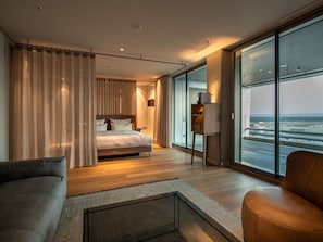 A modern hotel room with a large bed, a sofa, a small table, and a view of the airport. - b'mine Frankfurt Airport, WorldHotels Crafted (Frankfurt)