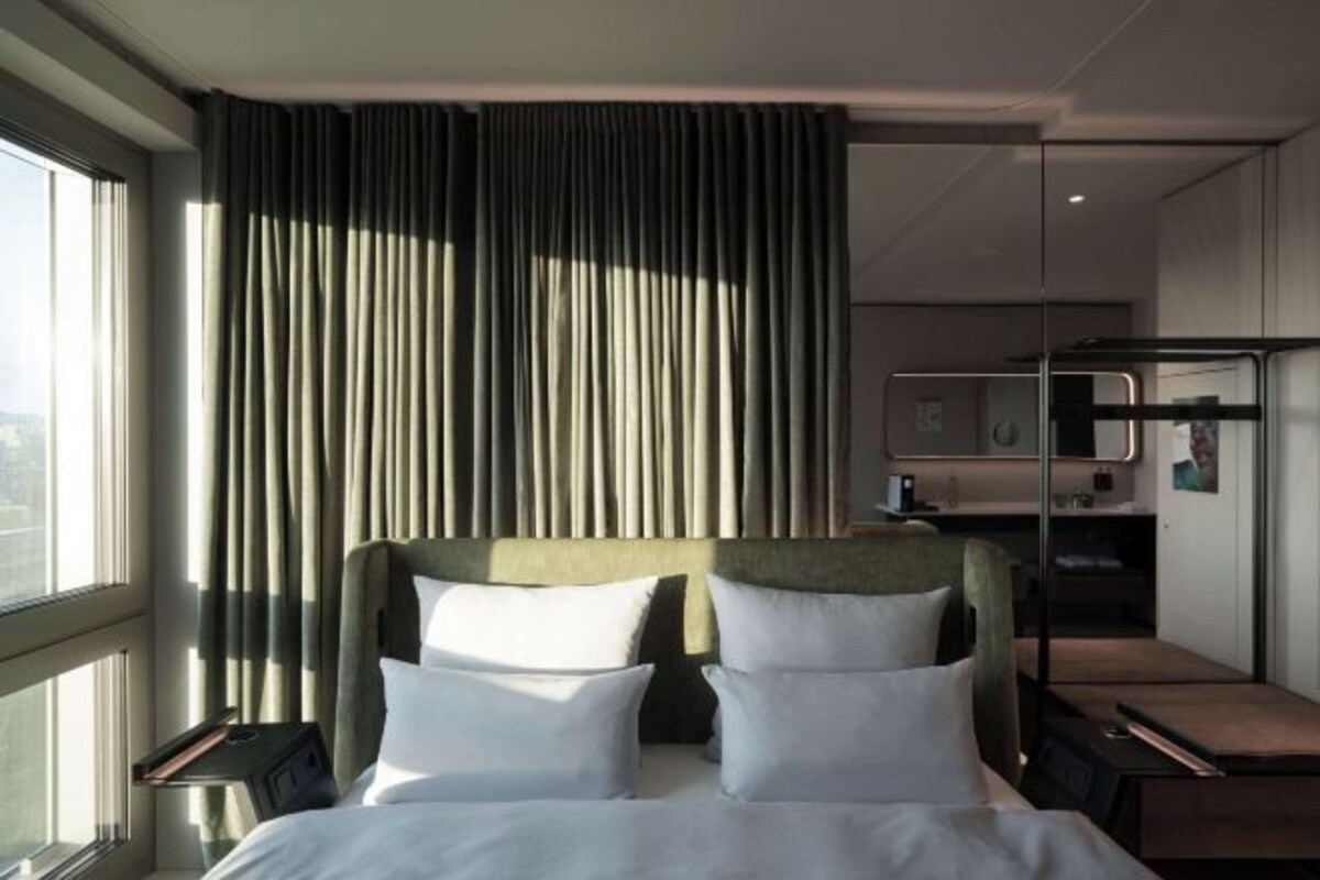 A modern hotel room with a large bed, a desk, and a view of the city through the window.