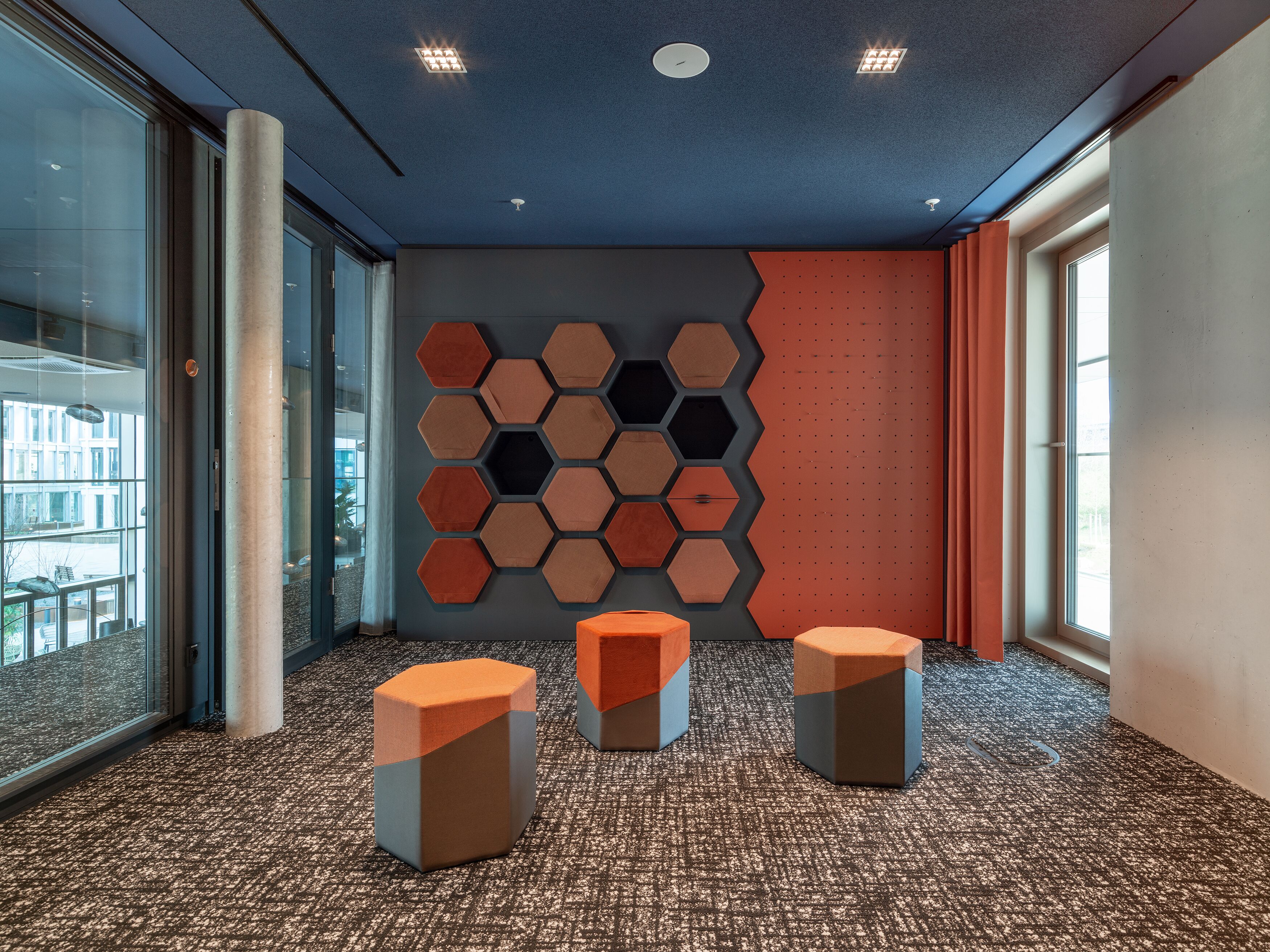 Un intérieur moderne avec des panneaux muraux hexagonaux, un sol recouvert de moquette et un mur en verre donnant sur l’extérieur.