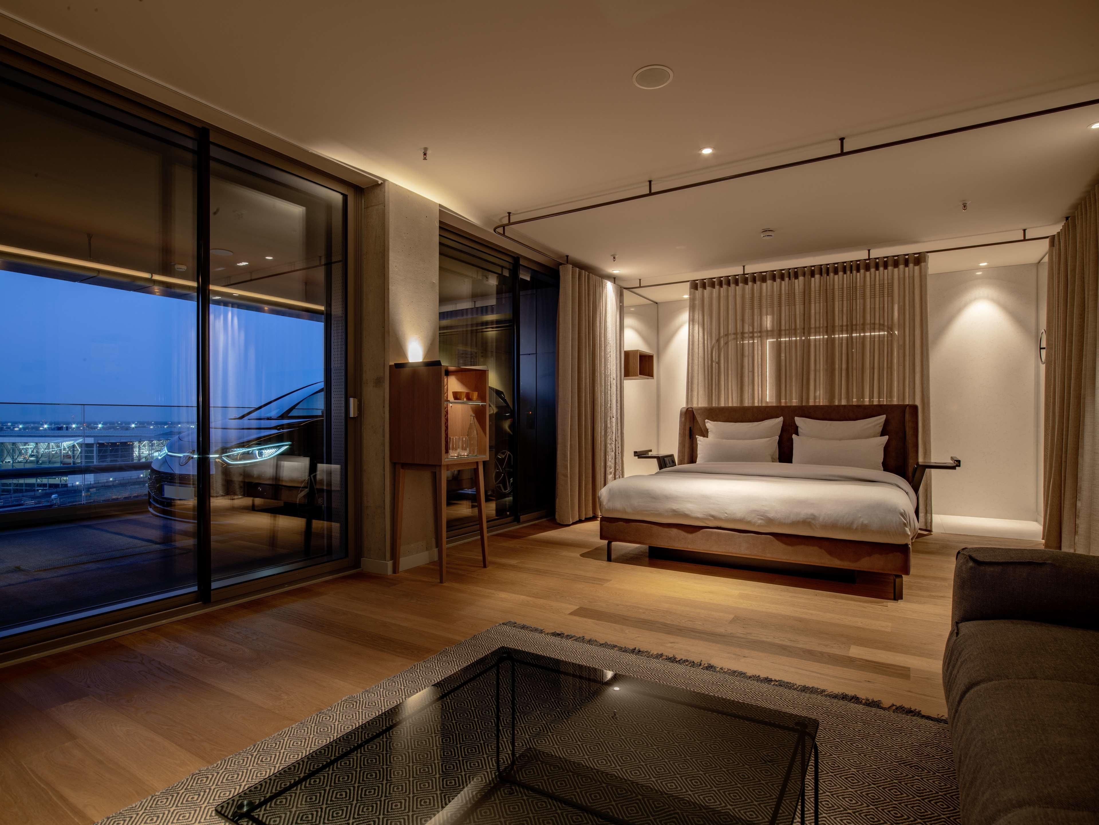 A modern bedroom with a large bed, a glass door leading to a balcony, and a small wooden side table.