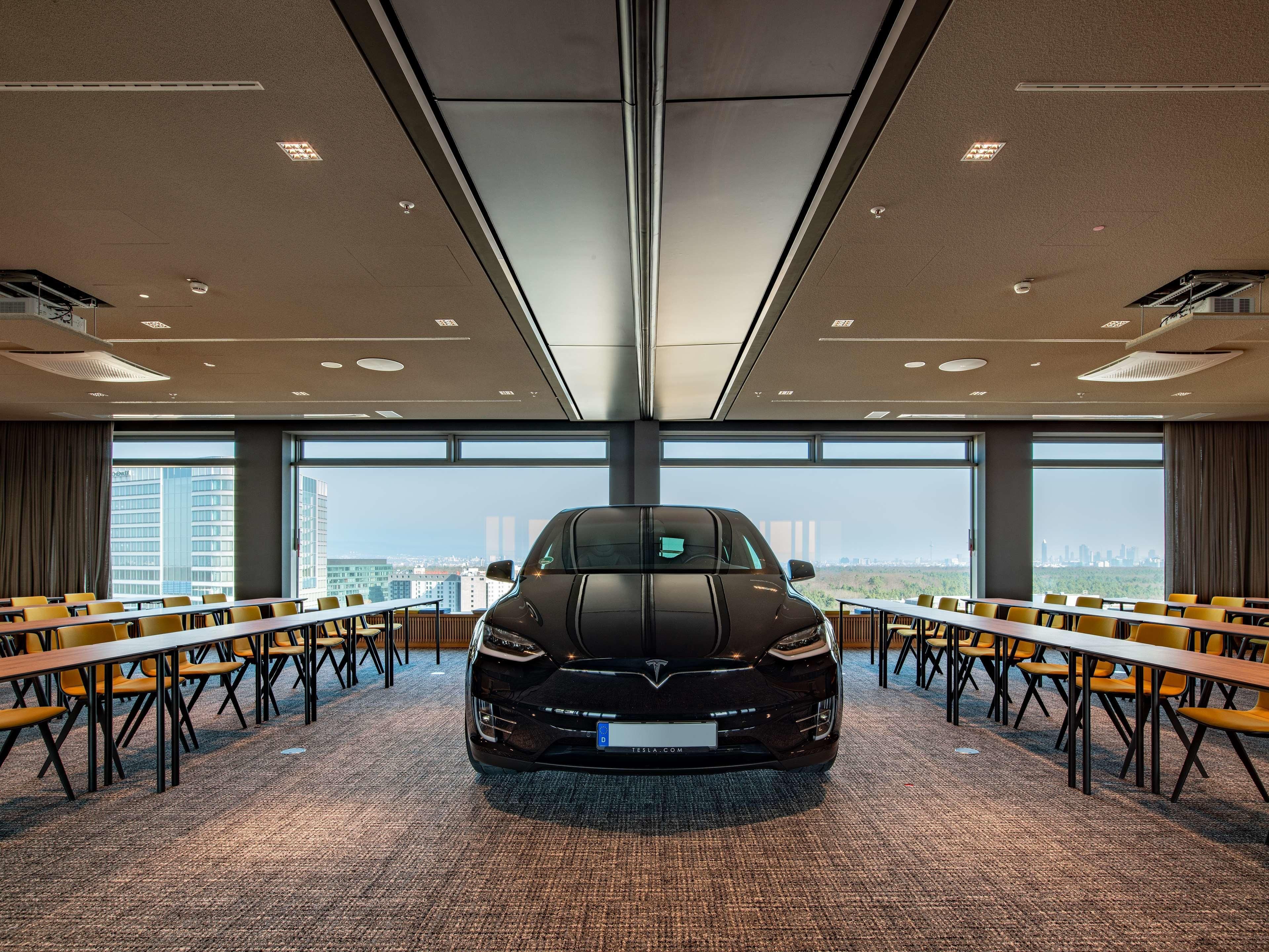 A conference room with a Tesla car parked in the center, large windows offering a city view, and tables with chairs arranged for meetings.