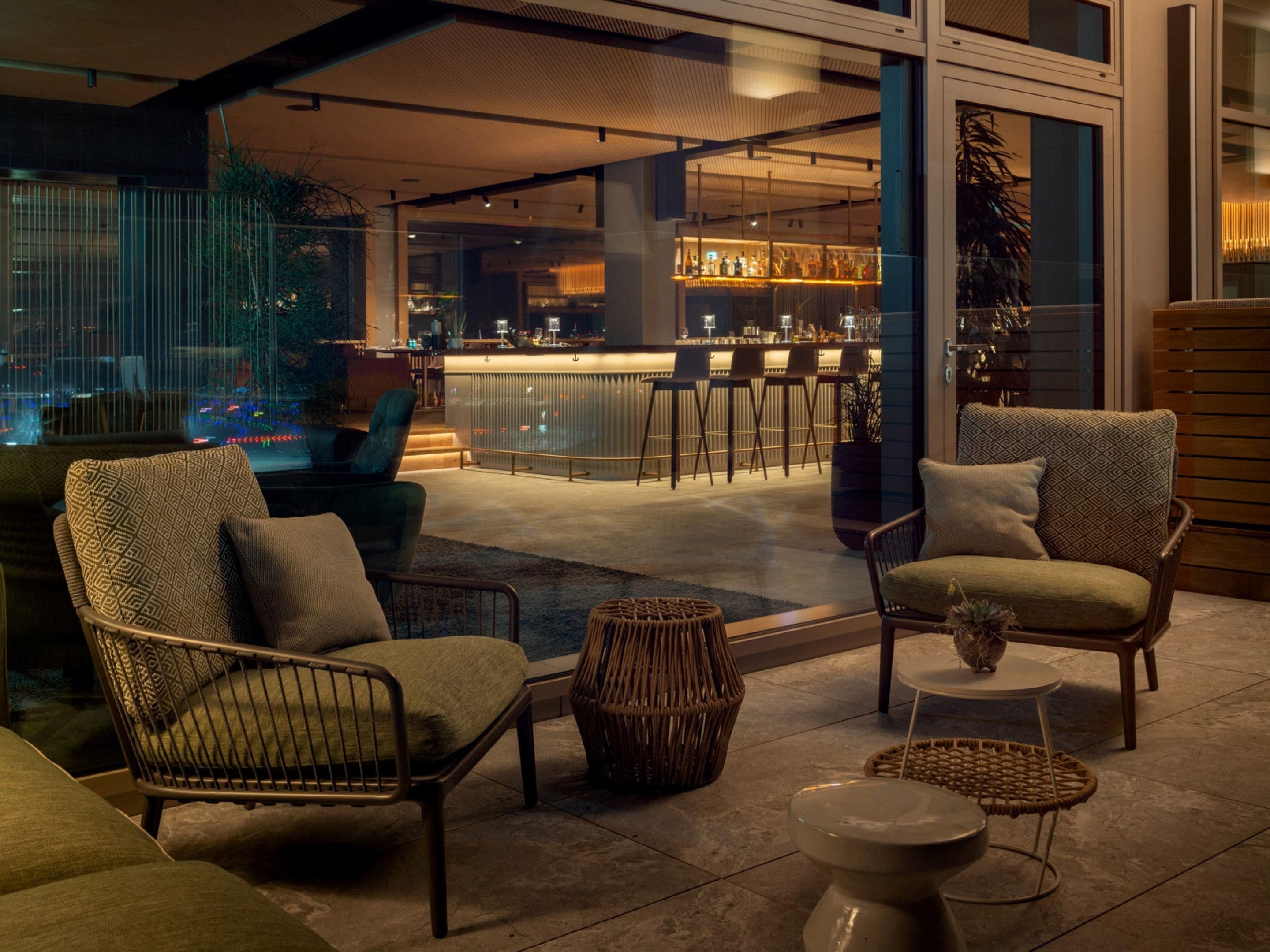 Un área de estar moderna al aire libre con mobiliario de mimbre, un bar al fondo y vistas de la ciudad.
