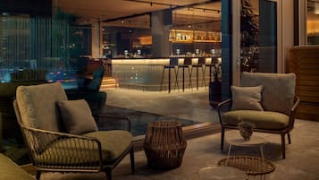 Un área de estar moderna al aire libre con mobiliario de mimbre, un bar al fondo y vistas de la ciudad.