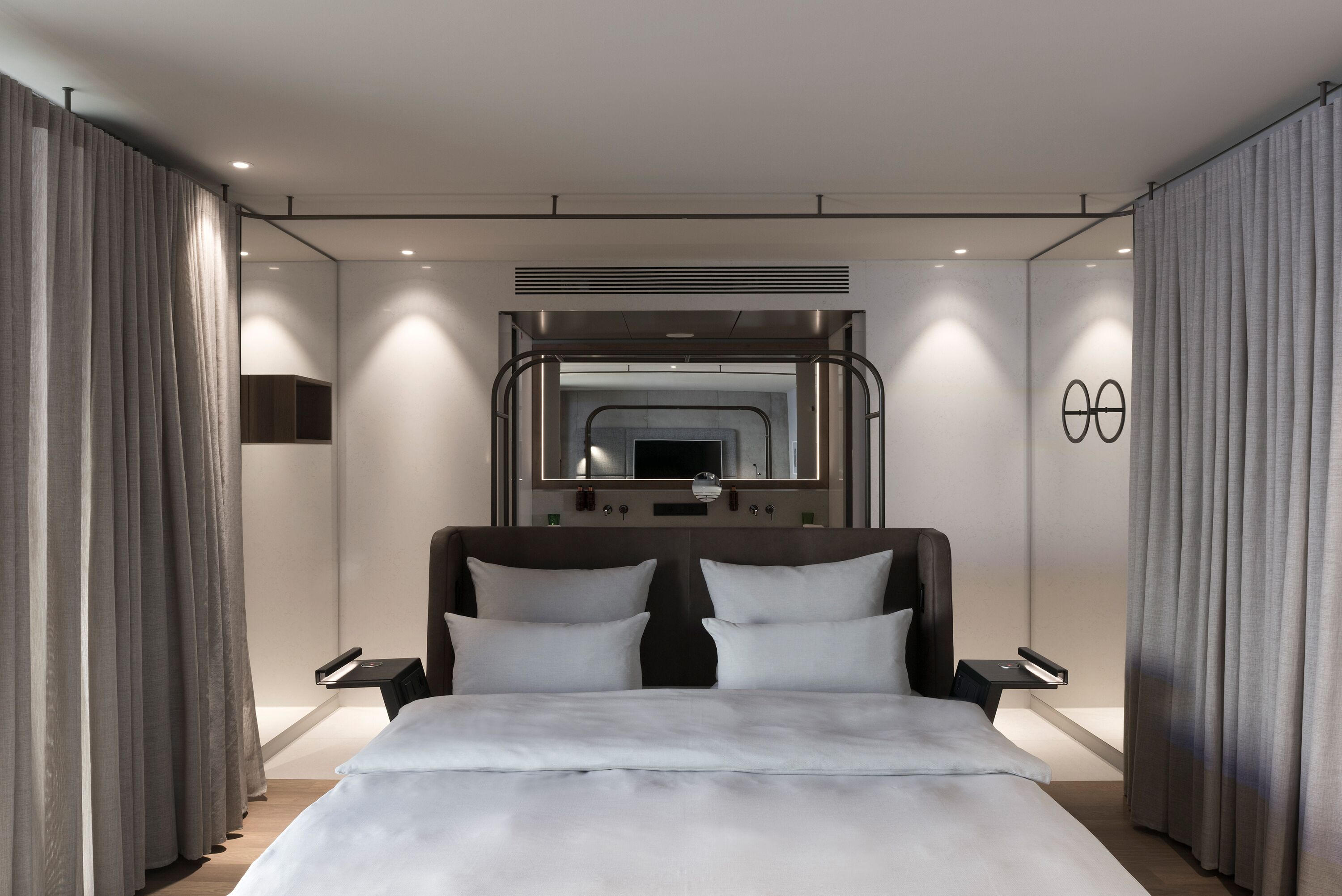 A modern hotel room with a large bed, a headboard with integrated lighting, a mirror, and a small bedside table.