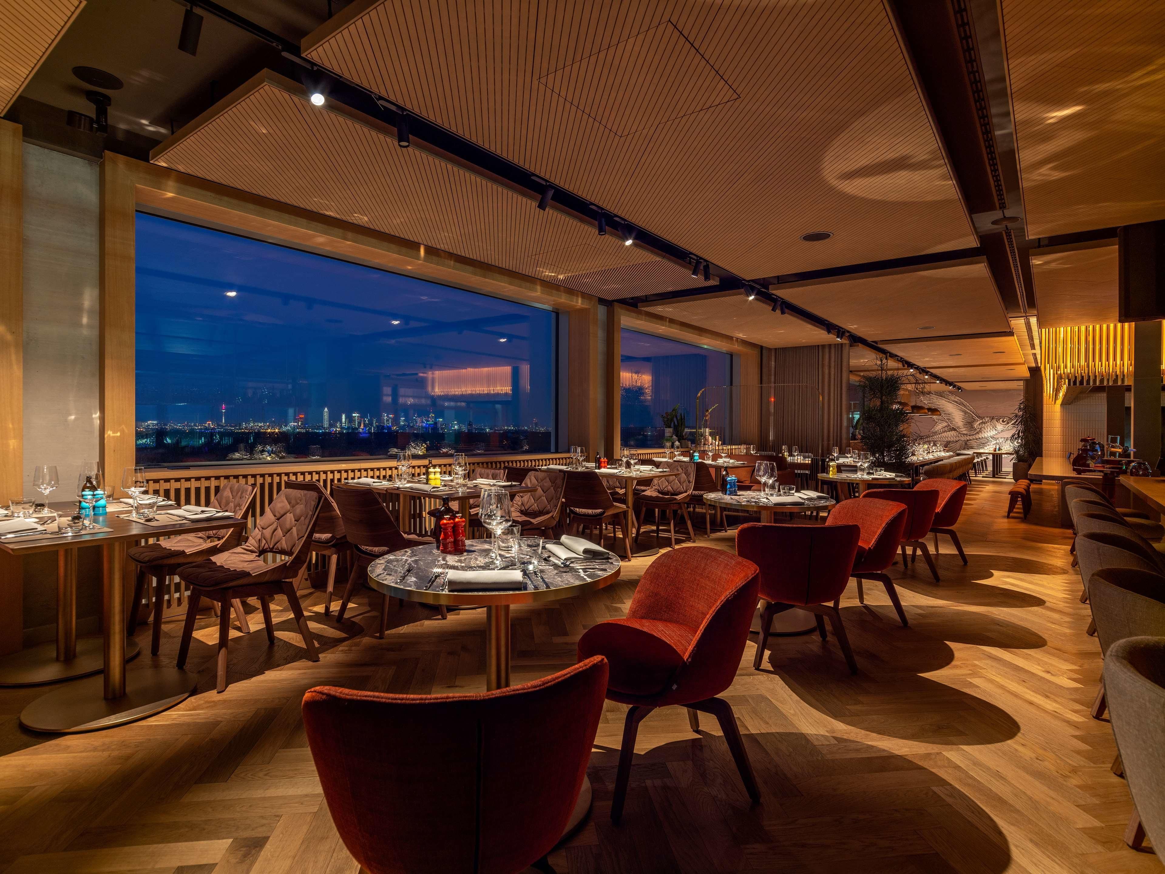 A restaurant with a view of a city skyline at dusk, featuring modern furniture and a wooden floor.