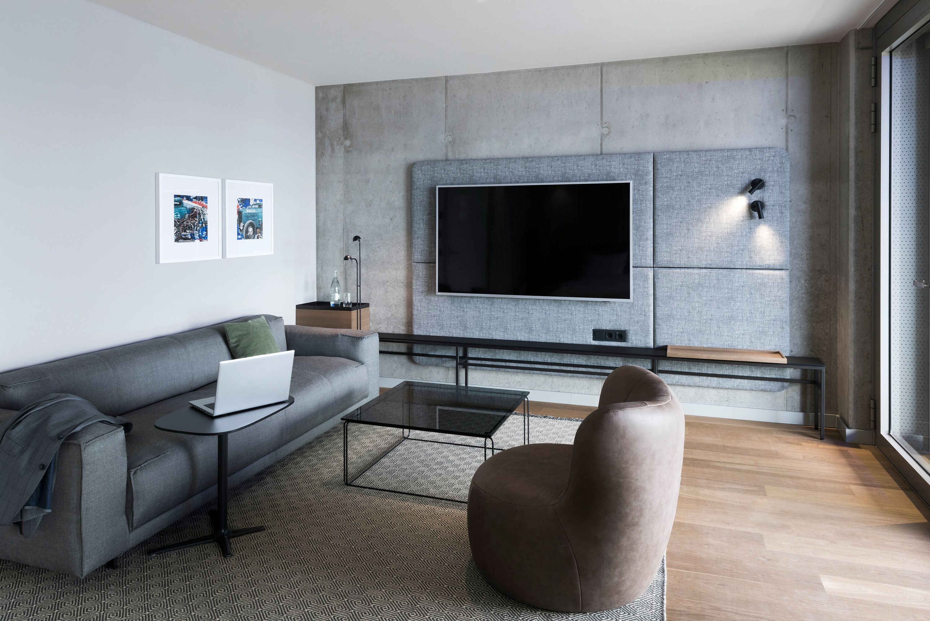 A modern living room with a grey sofa, a glass coffee table, a brown armchair, a flat-screen TV mounted on a textured wall, and artwork on the wall.