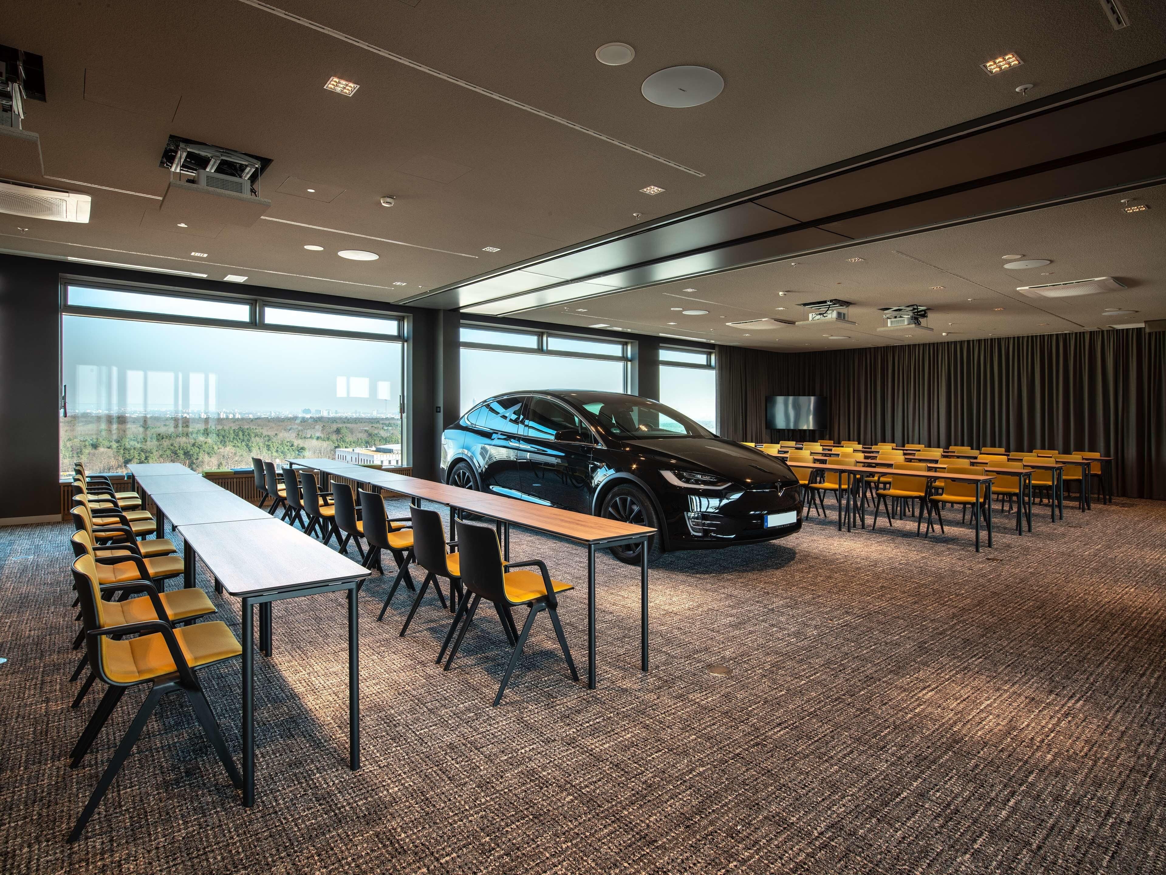 Een vergaderzaal met een lange tafel, stoelen, een auto en een groot raam.
