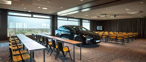 Een vergaderzaal met een lange tafel, stoelen, een auto en een groot raam.