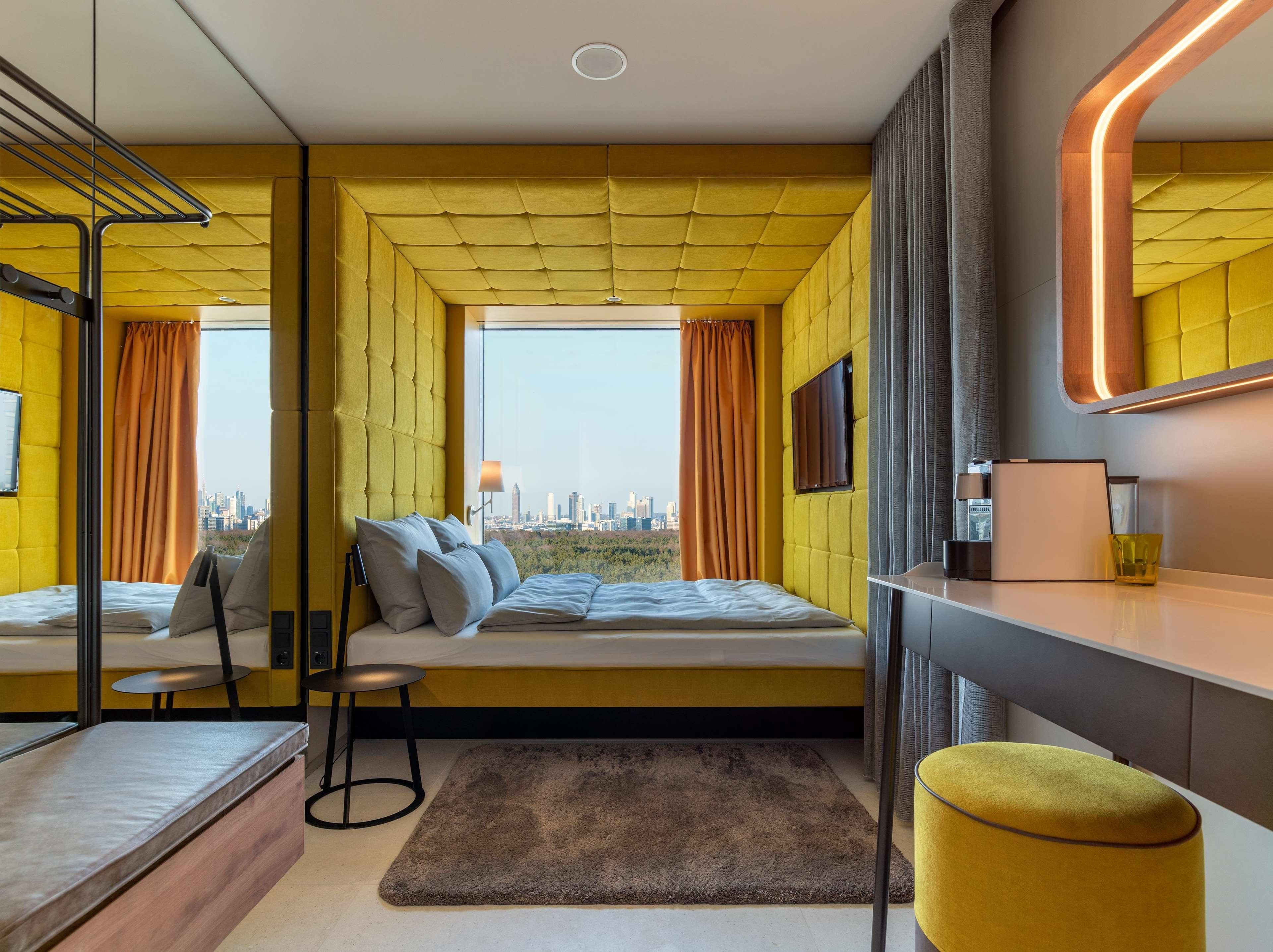Ein modernes Hotelzimmer mit einem Bett, einem kleinen Tisch, einem Stuhl, einem Schreibtisch und einem großen Fenster mit Blick auf die Stadt.