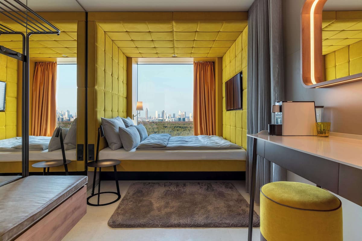 A modern bedroom with a city view, a bed with pillows, a small table, and a chair.
