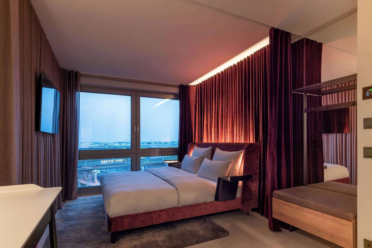 A hotel room with a large bed, a desk, and a view of the cityscape through floor-to-ceiling windows.