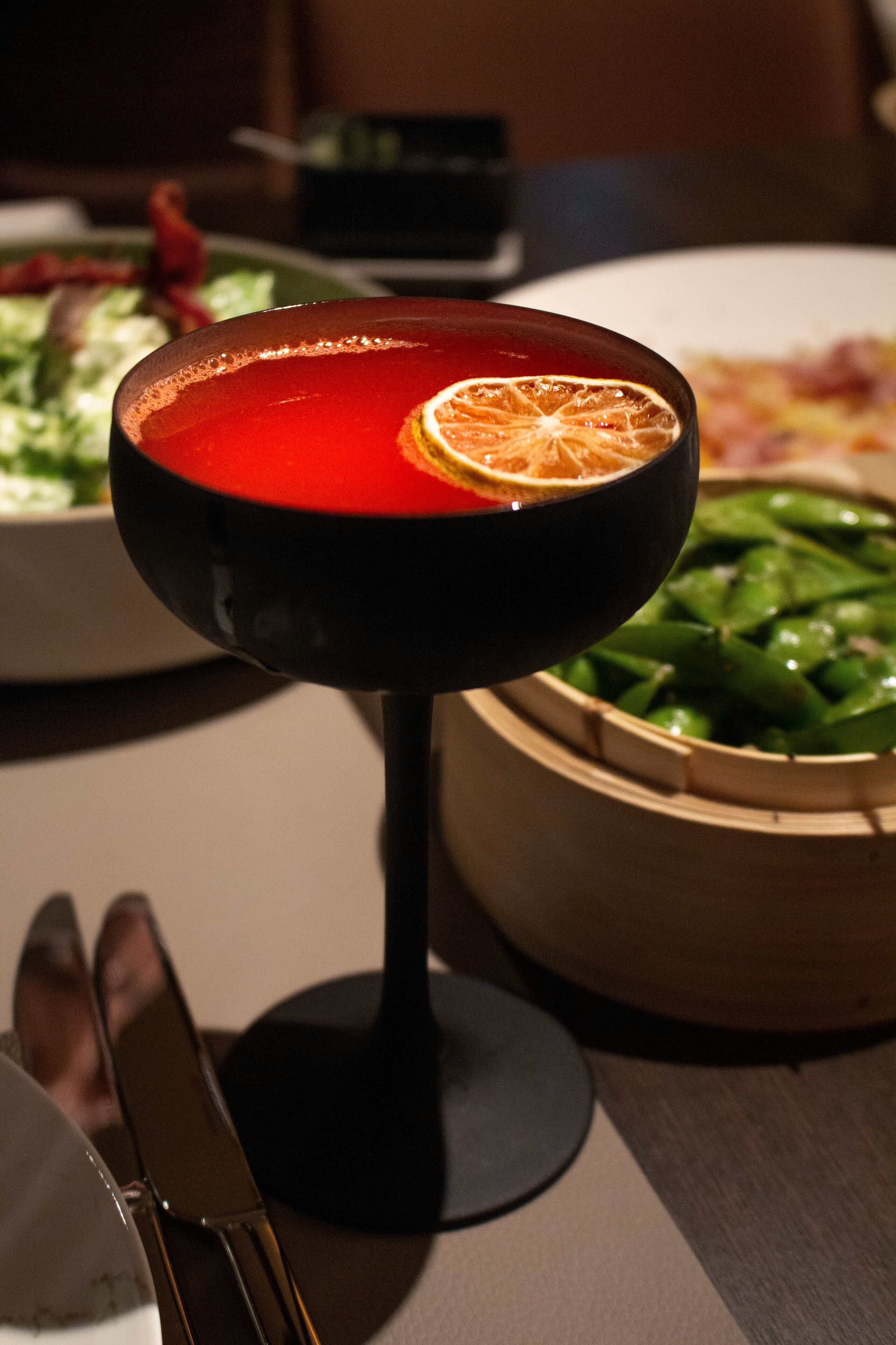 A table with a red drink in a glass, a bowl of green vegetables, and a plate of food in the background.