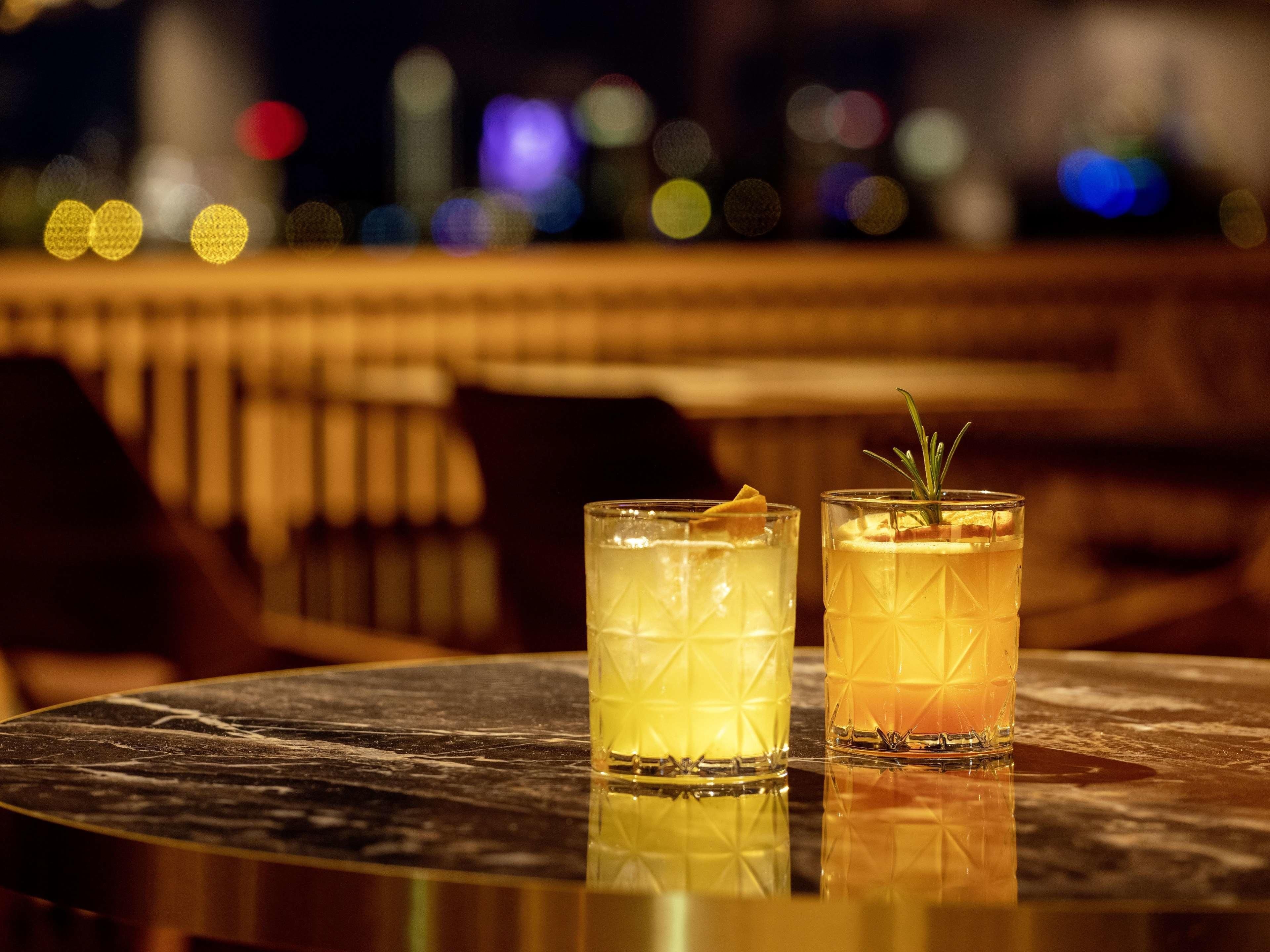 Dos vasos de una bebida amarilla sobre una mesa de mármol reflectante, con un fondo de paisaje urbano borroso.