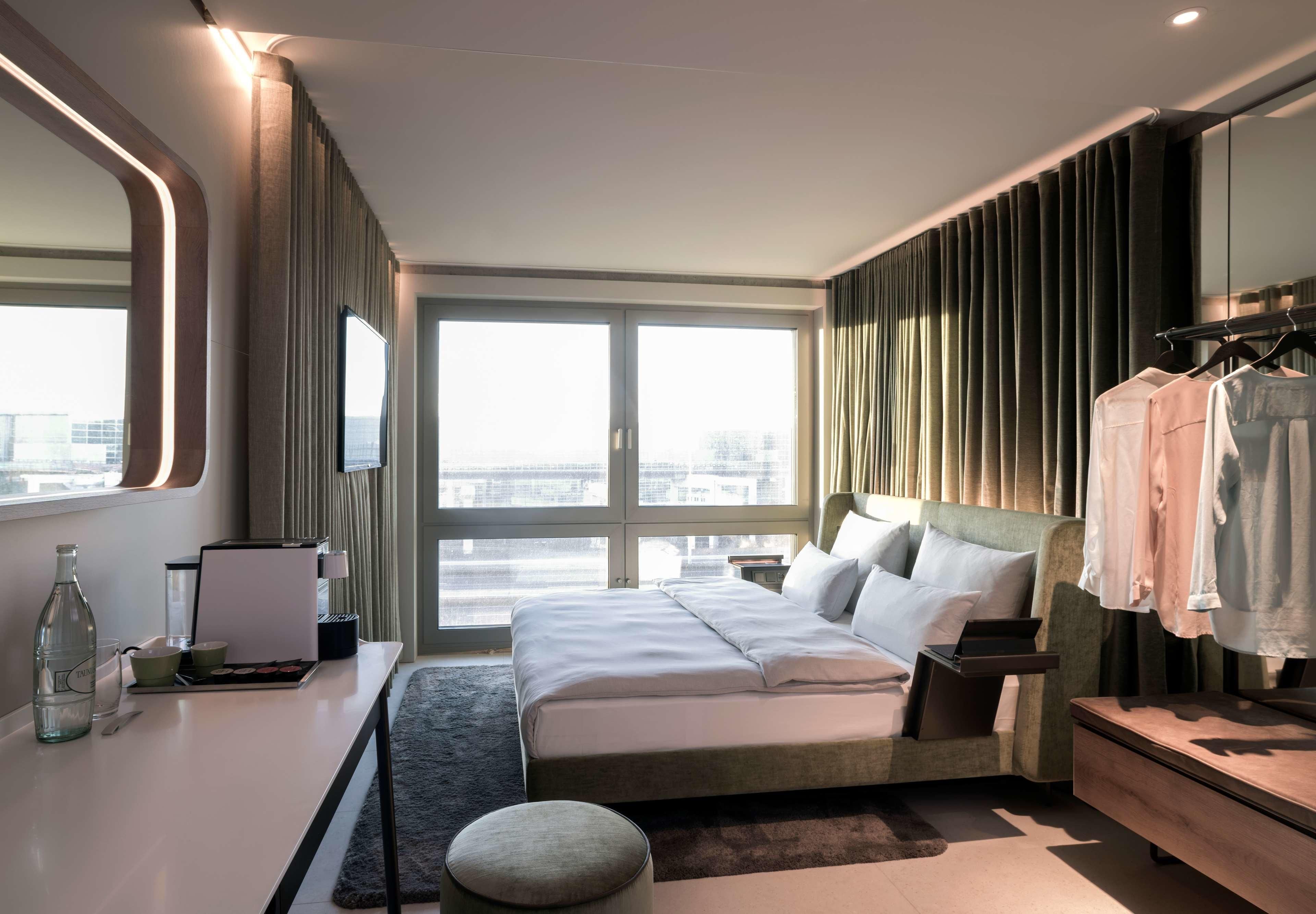 A modern hotel room with a large bed, a desk with a mirror, a vanity with a mirror, and a view of the city through large windows.
