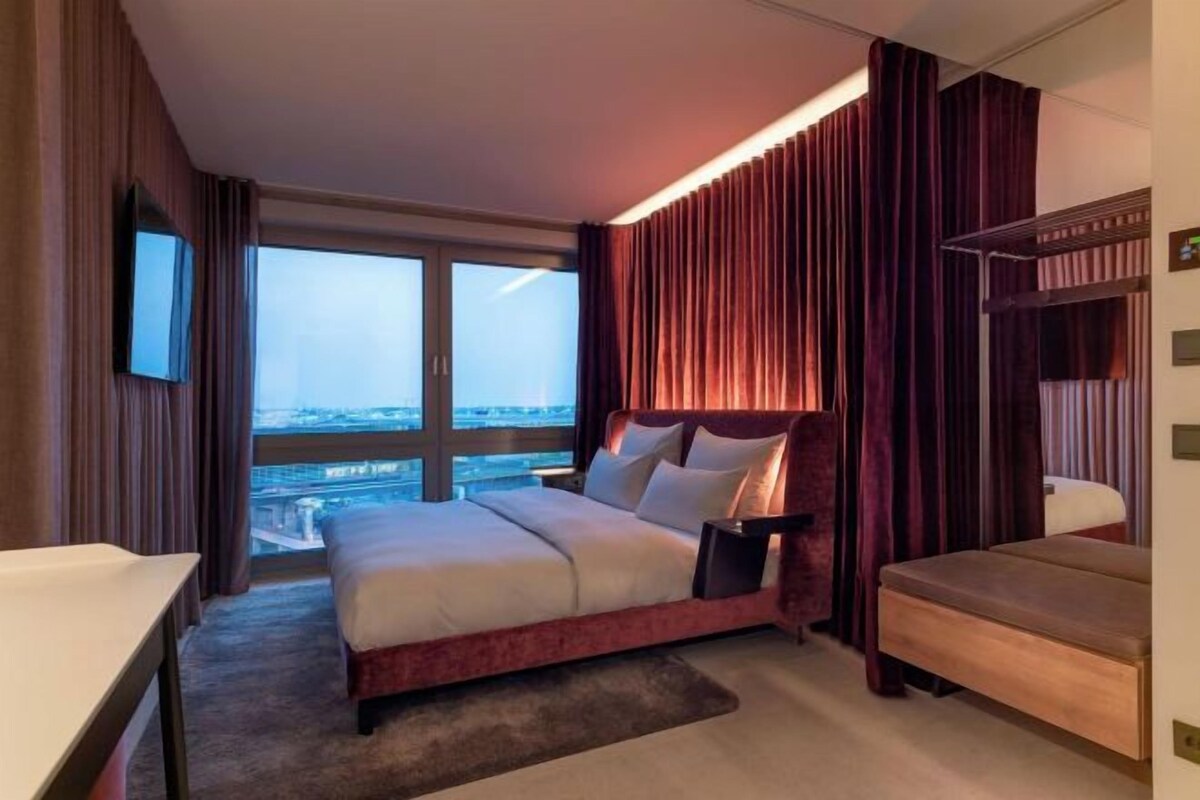 A hotel room with a large bed, a bedside table, a chair, a TV, and a view of the city.