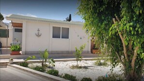 A single-story building with a flat roof, a small front porch, and a garden area with various plants and a tree. - Cottage a few steps from the sea (Gallina)