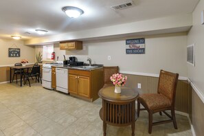 A compact kitchen with wooden cabinets, a stove, and a sink. There is a dining table with chairs and a small round table with a vase of flowers. A sign on the wall reads "WELCOME TO THE SHORE."