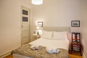 A neatly made bed with white linens and a patterned blanket, a bedside table with a lamp, and a framed picture on the wall.