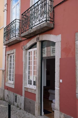 Um edifício com fachada vermelha, uma varanda com grades de ferro forjado e uma porta que dá para um quarto com uma cama.