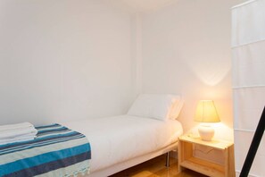 A single bed with a white comforter and a striped blanket, a bedside table with a lamp, and a wooden nightstand.