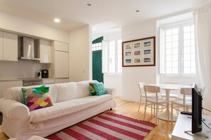 A modern living room with a white sofa, a red and white striped rug, a dining table with white chairs, a flat-screen TV, and a kitchen area with cabinets and a coffee machine.