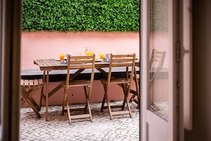Une table en bois, des chaises et un verre de jus d’orange, le tout placé devant un mur rose et agrémenté d’une haie verte.