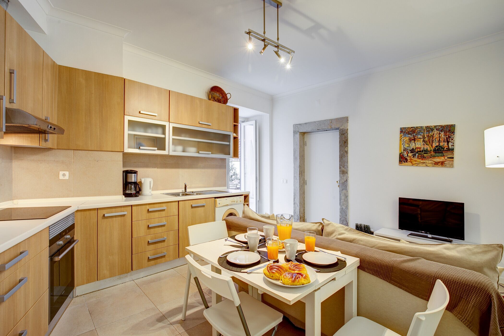 Een moderne keuken met houten kastjes, een eettafel gedekt voor twee personen en een televisie in de woonkamer.