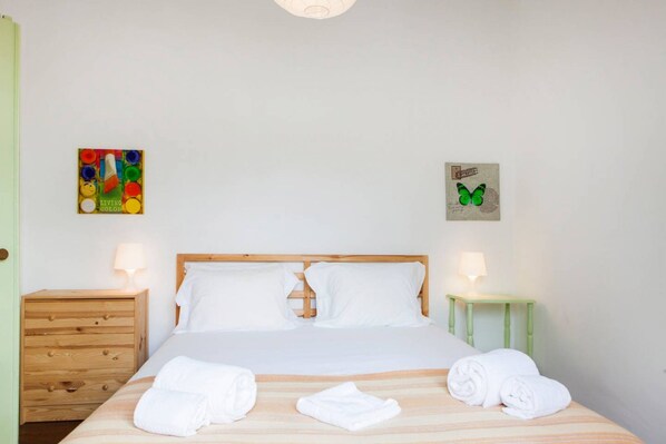 A neatly made bed with white linens and rolled towels, a wooden nightstand with a lamp, and a small green side table with a lamp.