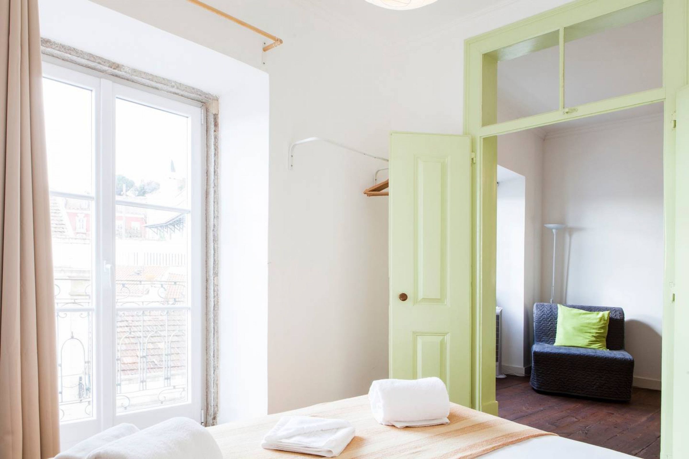 A room with a bed, a green door, a chair, and a view of buildings outside.