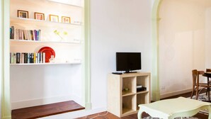 Una sala de estar con una estantería, un televisor sobre un mueble de madera, una mesa de comedor con sillas y una alfombra estampada.