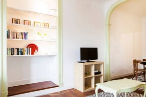 Un salon comprenant une bibliothèque, un téléviseur posé sur un meuble, une table à manger avec des chaises et un tapis à motifs.