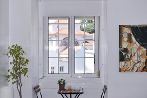 A room with a white wall, a window with a view of buildings, a table with a plant, and a painting.