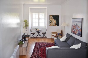 Una sala de estar con un sofá gris, un tapete rojo estampado, una pequeña mesa con sillas y dos cuadros en la pared.