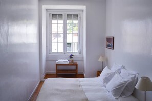 A bedroom with a bed, a bedside table with a lamp, a window with a view, and a small wooden bench with towels and a vase.