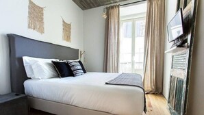 Ein Schlafzimmer mit Bett, Nachttisch, Wandfernseher und einem Fenster mit Vorhängen.